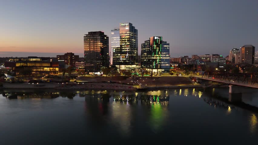 Downtown Saskatoon at dusk. River reflects city lights. Modern buildings glow against twilight sky. Bridge spans the water. Ideal for business or tourism content. Captures urban beauty from above