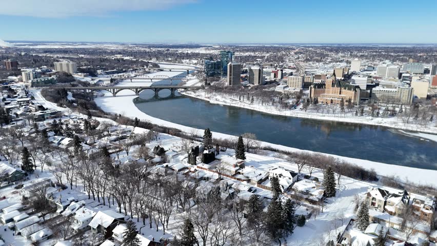 Aerial drone footage captures downtown Saskatoon in winter. Snow blankets streets and rooftops. Frozen river flows through the city. Bridge spans the river, connecting neighborhoods