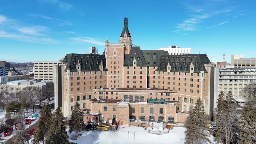 This drone video captures downtown Saskatoon. Historic building stands tall in winter. Snow blankets streets and trees. People walk calmly near the entrance. Clear blue sky enhances the scene