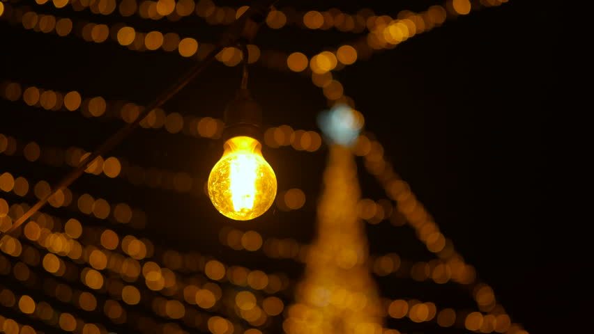 Golden christmas lights string glowing at night. Close up view of a glowing yellow light bulb swaying on a string. Blurry christmas lights and a decorated tree create a beautiful golden bokeh effect