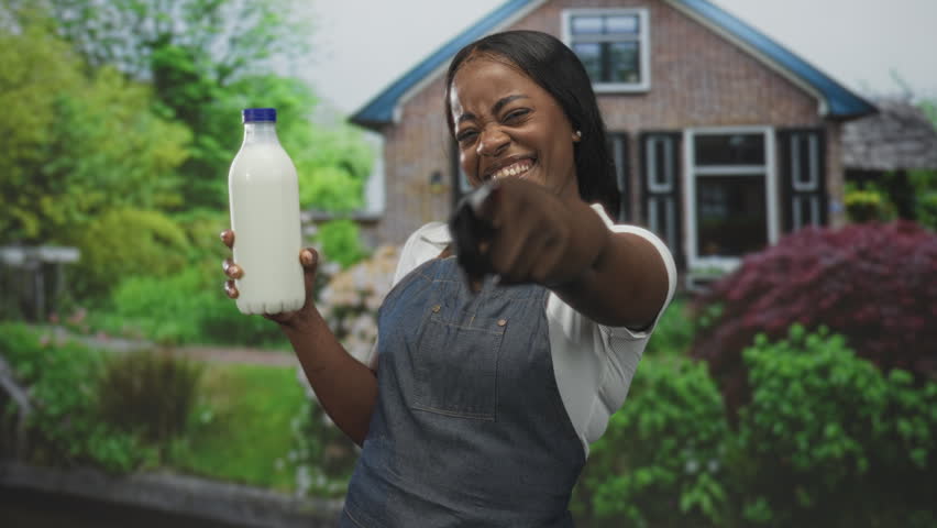 Woman wearing apron holds a milk bottle and points finger at camera in front of building; confidence playful charm.