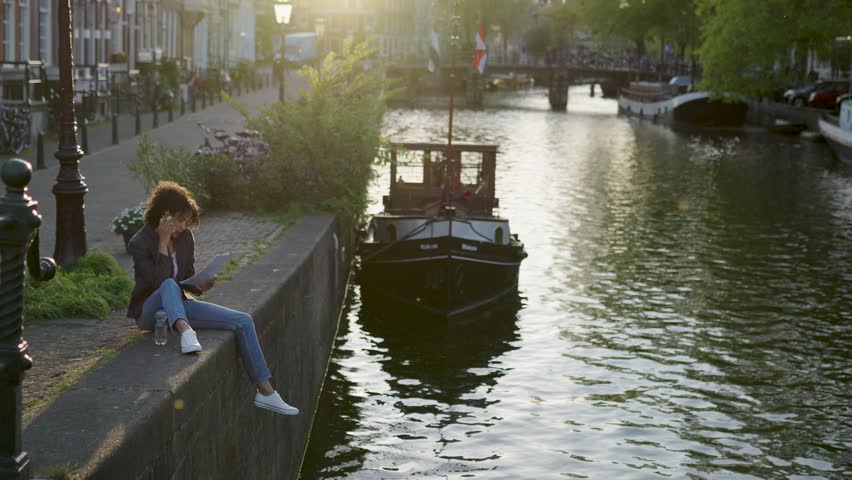 Beautiful mixed-race woman sitting by a canal in amsterdam, discussing business on her smartphone while holding documents at sunset