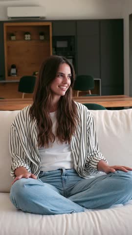 Smiling young woman enjoying a moment of relaxation on a comfortable sofa within a bright and modern home interior. Her joyful expression adds warmth to the domestic scene.