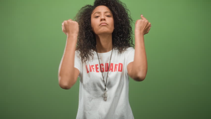 Young woman with curly hair shows middle fingers and whistle while wearing lifeguard shirt in studio green backdrop; defiance attitude.