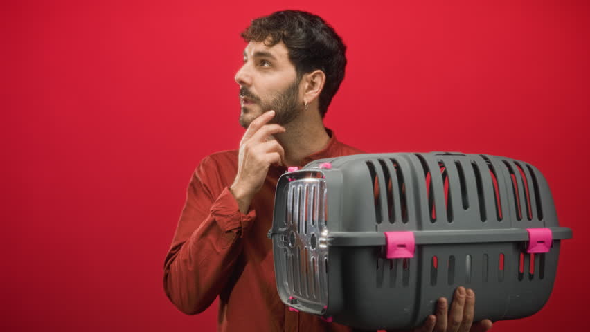 Man holding pet carrier with one hand under its base and other hand to chin while looking upward in studio; quiet contemplation.