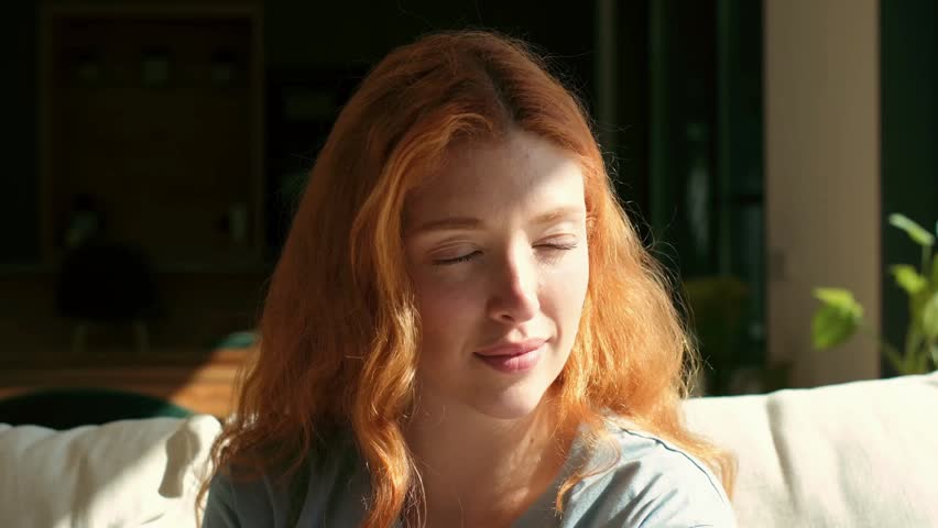 A beautiful young woman with long red hair relaxes on a sofa at home, gently closing and opening her eyes, enjoying the warm sunlight on her face in a moment of peace and tranquility.