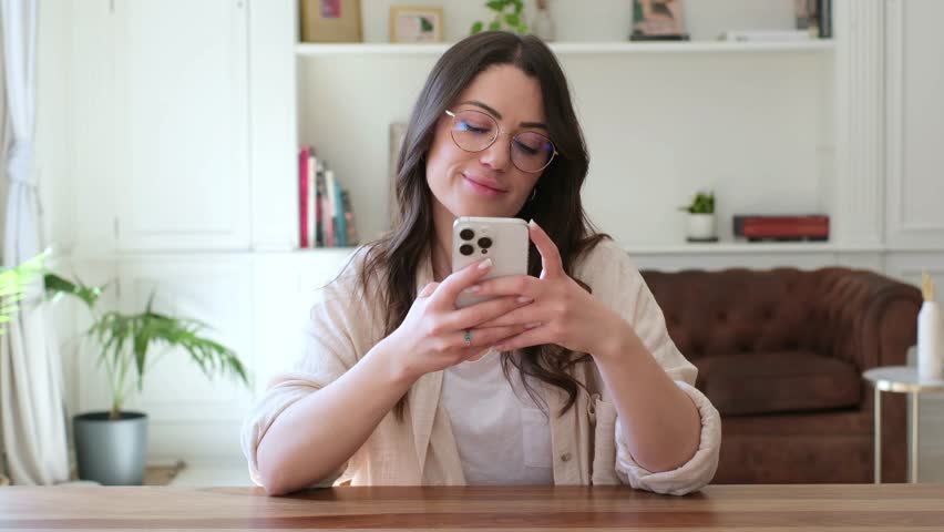A happy young woman with stylish glasses holds her cell phone, smiling and looking up thoughtfully. She enjoys a peaceful moment, perhaps browsing or connecting online in her modern home interior.
