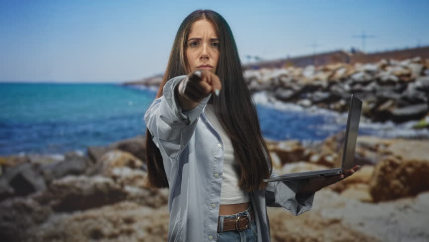 Young hispanic woman holds laptop and beckons with outstretched hand at rocky beach shoreline, stern expression visible; determination challenge.