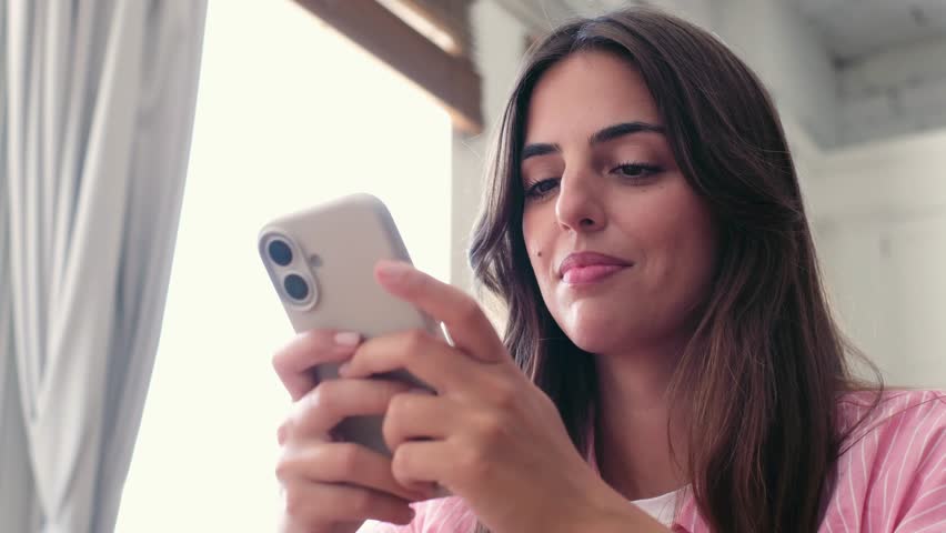 A happy young woman is seen smiling brightly while interacting with her mobile phone. She appears content as she enjoys online communication and social media from the comfort of her home.
