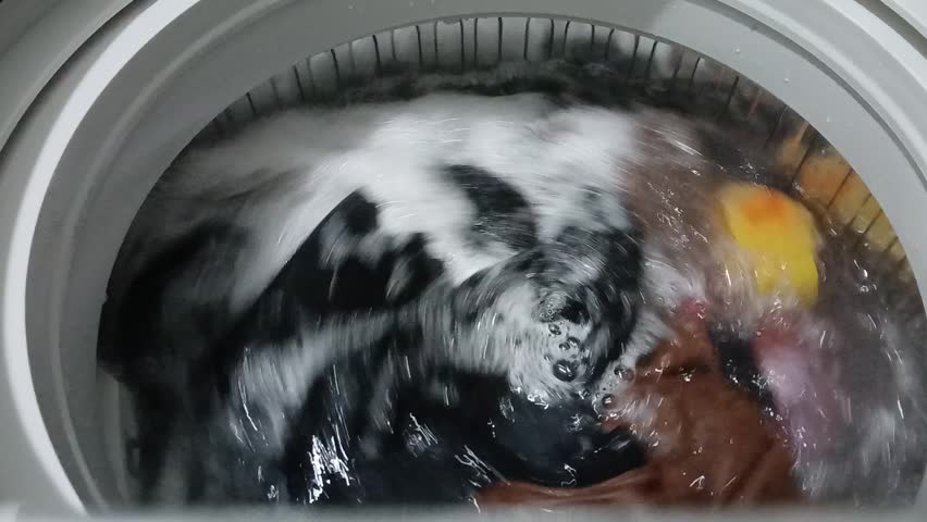 Top View of Washing Machine Agitating Clothes with Soap Suds and Water. A high-angle, close-up shot looking down into a top-loading washing machine during its wash cycle. The video captures the powerful circular motion of the water and colorful laundry, including black, blue, and brown garments, spinning through white soap suds and bubbles. The clear water and mechanical agitation create a sense of cleanliness and domestic routine. The clip includes the original mechanical rhythmic sound of the appliance. Perfect for content related to housekeeping, laundry services, home appliances, and daily lifestyle chores.