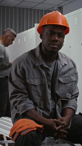 African American Factory Worker Sits on Bench, Wearing Orange Safety Helmet, Grey Jacket, With Metal Lockers and Colleagues in Background. Industrial Camaraderie, Diversity. Portrait. Vertical Shot.