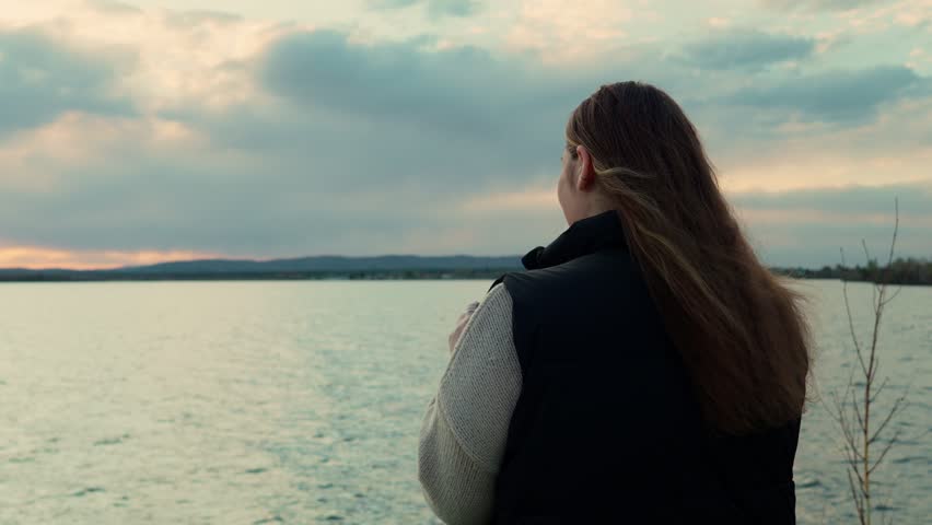 A girl is standing on the shore of a lake, looking through binoculars and admiring nature. The weather is overcast and cloudy.