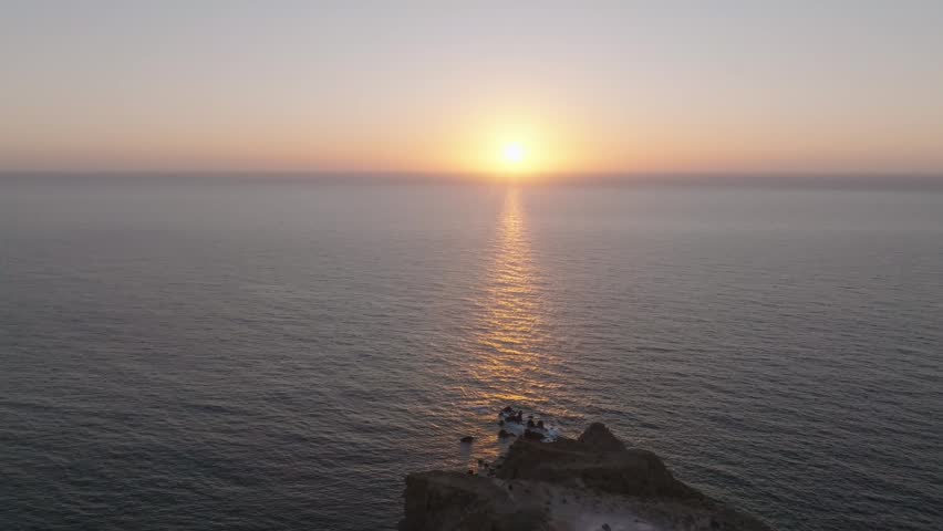 Drone pullback footage of the sun setting on the Santorini horizon, revealing the lighthouse as a perfect spot to watch the sunset over the Aegean Sea.