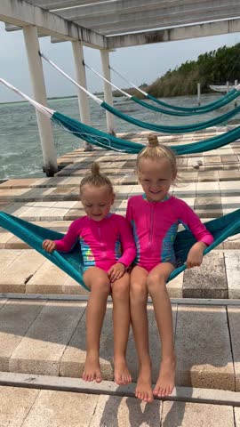 Two little sisters in matching swimsuits relaxing together on a hammock by a tropical lagoon, happy family vacation. Vertical video