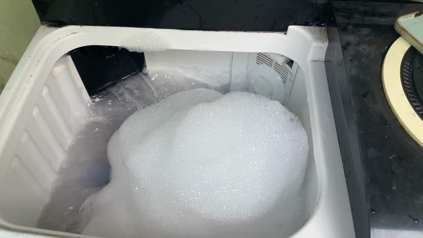 Close-up of a washing machine tub overflowing with a massive amount of thick, white soap suds and bubbles due to too much detergent or a malfunction.