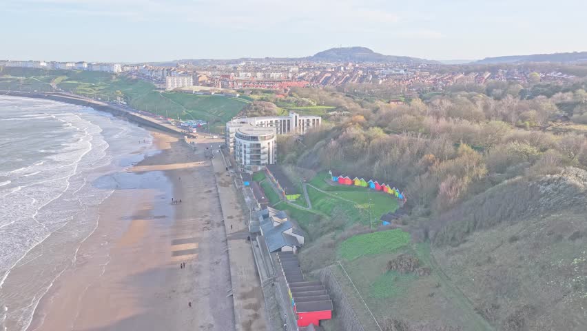 North Bay beach in Scarborough, North Yorkshire, England, United Kingdom, with curved shoreline, promenade, hotel buildings, woodland hillside and colorful beach huts under soft daylight drone