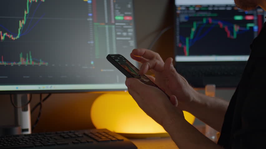 Man checking trading app on smartphone. Trader using mobile phone at desk with stock market charts on monitors. Concept of online trading, financial analysis, investment and digital finance