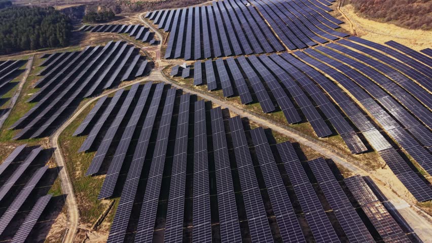 Solar panels cover a large area in a field. Rows of panels reflect sunlight. Green grass and trees surround the field. The scene shows technology for renewable energy production.