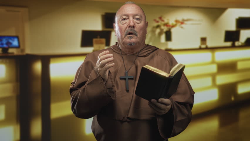 Man monk holding bible and clutching wooden cross pendant with right hand in building reception area; devotion.