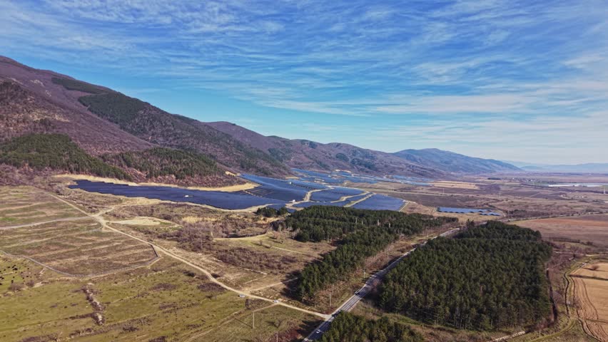 Solar panels stretch across the landscape next to large areas of trees and mountains. The sky shows clear weather, and the sunlight illuminates the scene with a blue backdrop.