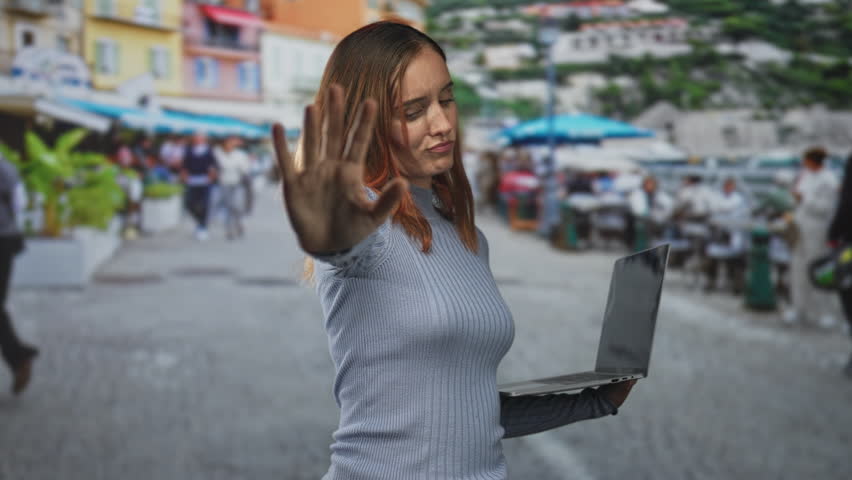 Woman redhead holding laptop and showing palm raised to signal stop on a street crowded promenade; defiance privacy.