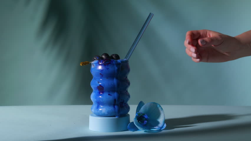 Dynamic push in shot of female hand grabbing vibrant blue spirulina cocktail topped with blueberries gold garnish and glass straw on teal
