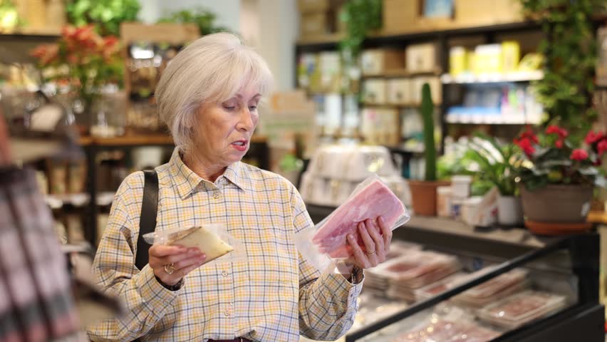 Mature woman with gray hair in an eco supermarket chooses a package of cheese and bacon. Senior woman buys groceries from organic store. High quality 4k footage