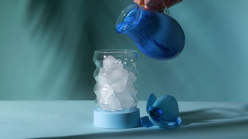 Vivid blue spirulina matcha milk pouring from pitcher over crystal ice into wavy bubble glass beside orchid on teal surface in slow motion