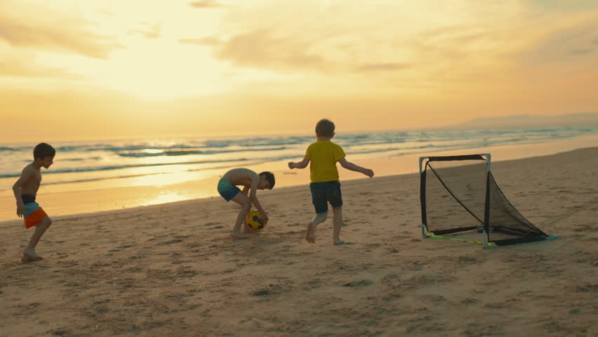 Happy kids running and playing football on a sandy beach by the sea during a beautiful summer sunset