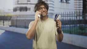 Man holding creditcard and phone to ear, smiling while talking and displaying cards near a stone building and iron fence on a street; confidence convenience. - Powered by Shutterstock - Get 15% off with code: PIKWIZARD15