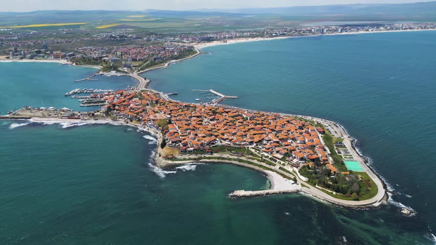 High angle drone shot of the historic Old Town of Nessebar, Bulgaria. A UNESCO World Heritage site situated on a scenic peninsula surrounded by turquoise sea water under bright sunlight.