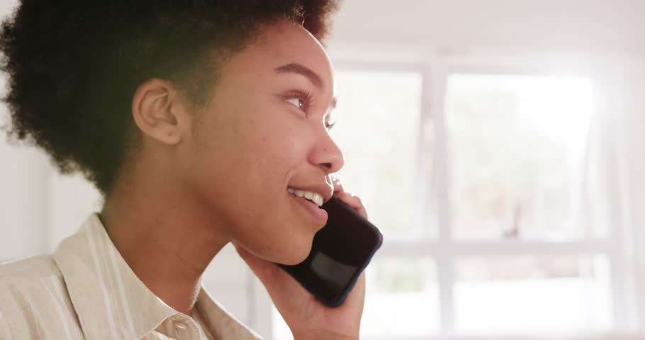 African American woman holding phone smiling speaking at home by window, light shirt, copy space. Smartphone, call, cheerful, backlit, sunlit, striped, collared