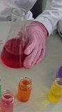 A person in a lab coat pours red liquid from a flask into a beaker. Several small containers with different colored liquids are on the table. This takes place in a lab setting during a science class. - Powered by Shutterstock - Get 15% off with code: PIKWIZARD15