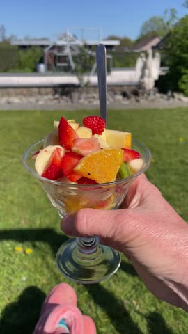 Close-up of a hand holding a glass bowl filled with fresh fruit salad on a sunny garden background,Healthy summer snack with strawberries, oranges, and apples on green grass lawn