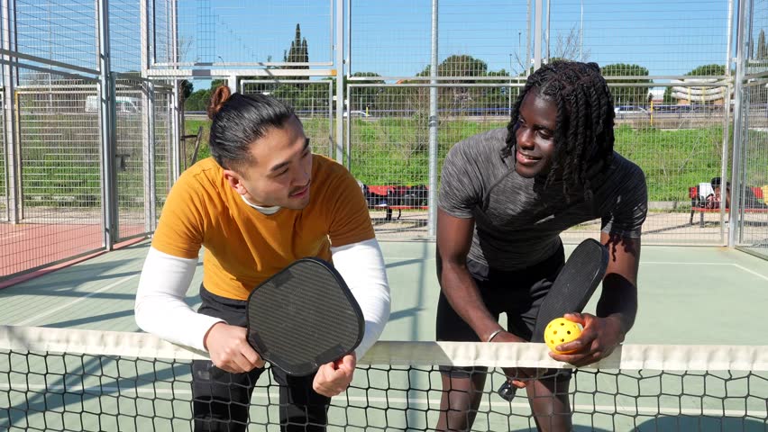 Two young multiethnic men standing by the net on an outdoor court, holding paddles and a ball while talking and laughing together after a friendly pickleball game on a sunny day