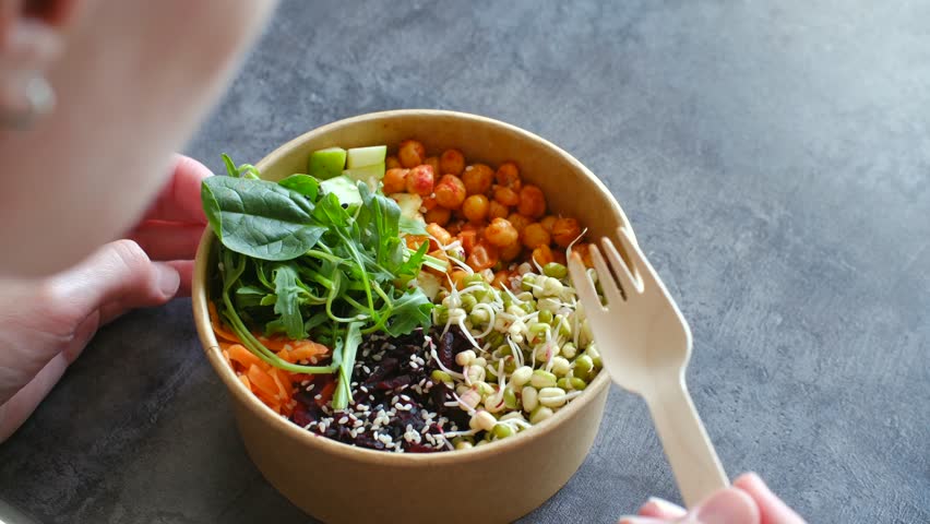Woman hands holding a takeaway kraft paper bowl filled with healthy vegan meal of chickpeas, sprouts, and fresh greens, eating with a wooden fork over gray concrete background. Healthy eating concept.