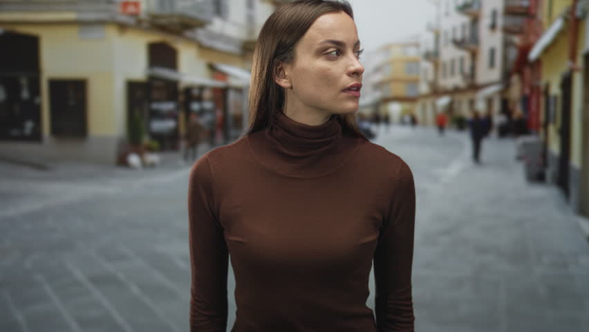 Young woman stands looking to the side, face visible, wearing brown turtleneck on a cobblestone city street with blurred storefronts and pedestrians; contemplation.