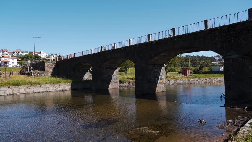 A close-up still of Ponte Grande da Torna in Ribeira de Oleiros shows the bridge standing firmly above a stream of crystal-clear water, with a bright blue sky stretching wide behind it. 4K