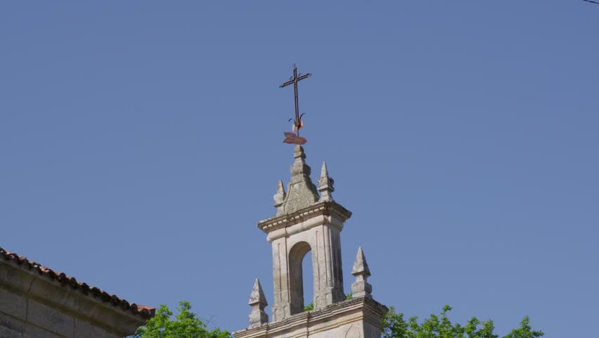 A static medium shot of an ornate stone church steeple topped with a cross, set against a vibrant blue sky and lush green treetops in bright sunlight.