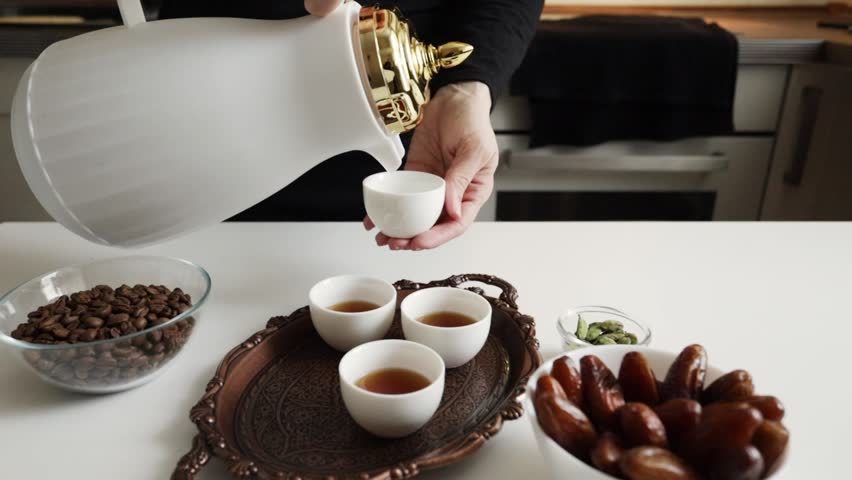 arabic coffee being poured from white dallah into a finjan cup on a copper serving tray with filled cups, dates and cardamom on the table