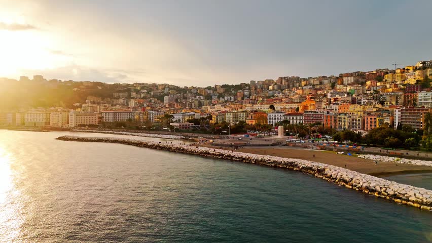 Aerial view of Naples, Italy, showing the curved waterfront barrier, public beach edge, and densely built hillside neighborhoods in warm late-day coastal light.