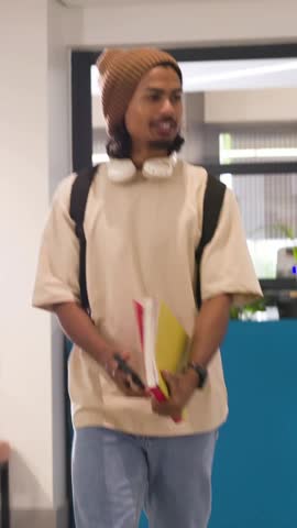 Vertical video: Entering man with pack holding red file in lounge, placing it to study on teal roll. Coworking, student, workspace, modern, organized, productive, vertical video