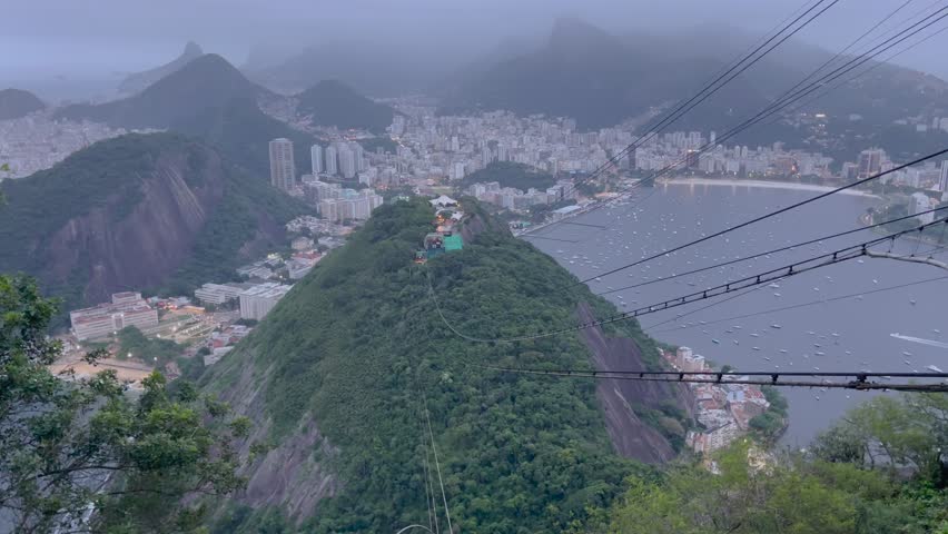 Static aerial view from Sugarloaf Mountain at dusk, cable car wires foreground, Botafogo Bay, misty mountains, Rio de Janeiro cityscape.