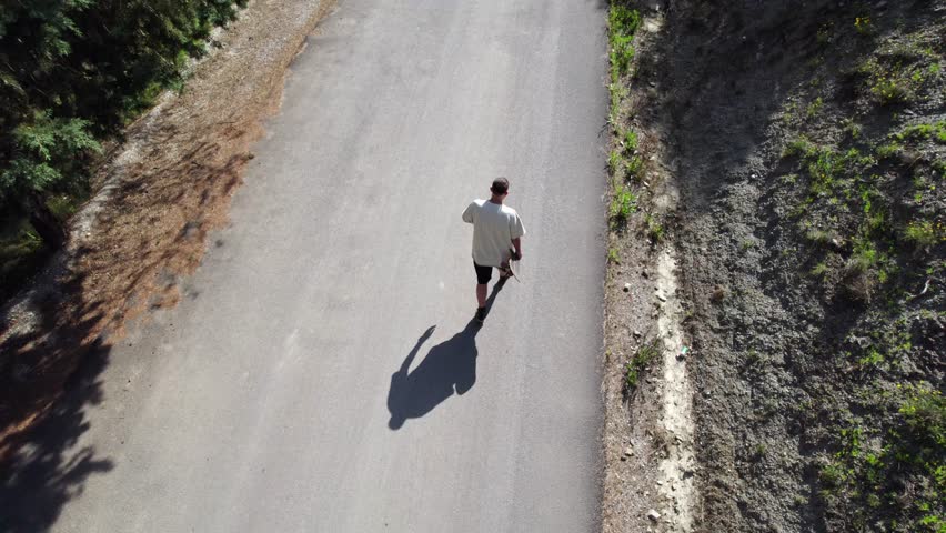Aerial view of a young man carrying his cruiser skateboard while walking down a shady road surrounded by a pine forest on a sunny day