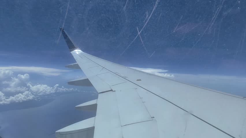 View of an airplane wing seen from a passenger window while flying above the clouds under a clear blue sky. A travel and aviation concept with a wide open sky, aerial perspective, and in-flight window view