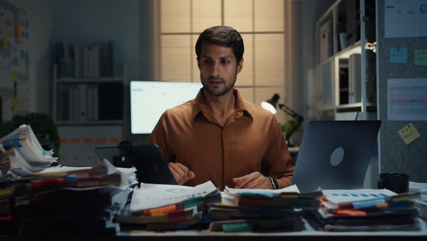 Overworked young Latin businessman working late on document in large stack of paperwork files at office night. Tax audit deadline, accounting stress, corporate burnout, urgent business concept.