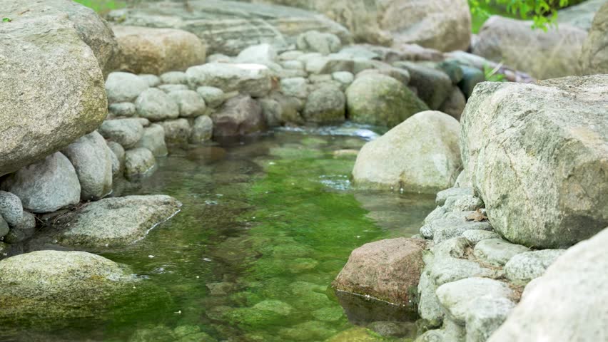 Clear, calm stream flows gently among large, smooth stones. Water reflects soft green moss growing on rocks. Scene feels serene and untouched by human activity