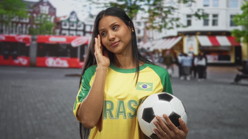 Woman in yellow brazil football jersey holds a soccer ball and wipes her face with one hand while standing on a crowded city street; disappointment.