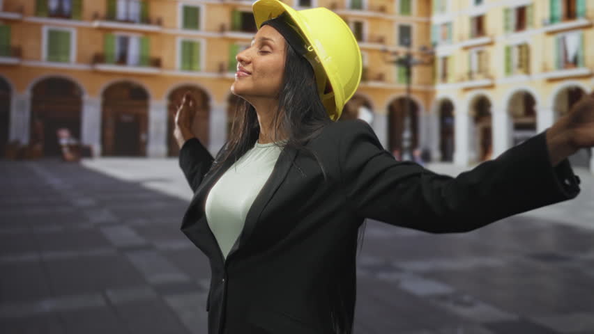 Woman wearing yellow hardhat smiles with eyes closed and tilts head back on street plaza with arched colonnades, blazer on; confidence progress.