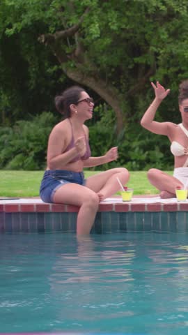 Vertical video: Talking woman in purple bikini splashing foot at home pool teasing friend with cup. Friends, backyard, outdoor, sunny, casual, laughter, vertical video